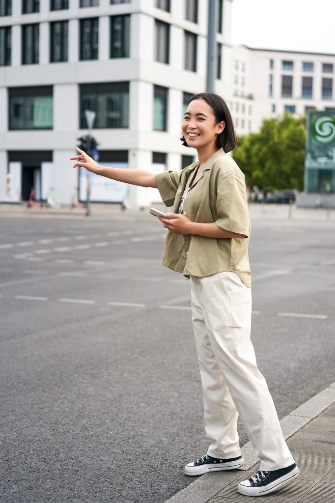 Vertical shot of young woman trying catch taxi, waving at driver on road, holding smartphone with